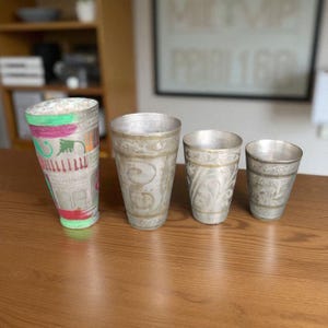 May include: Four silver-colored drinking glasses of varying sizes. One glass has a colorful design with pink, green, and orange accents. The other three glasses have ornate, etched patterns and a metallic finish.