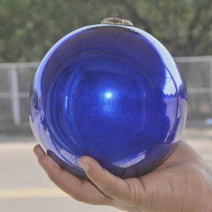 Puede incluir: Un adorno navideño grande y redondo de vidrio azul cobalto con una superficie brillante y reflectante. El adorno tiene una pequeña tapa metálica en la parte superior para colgarlo. El fondo está borroso.