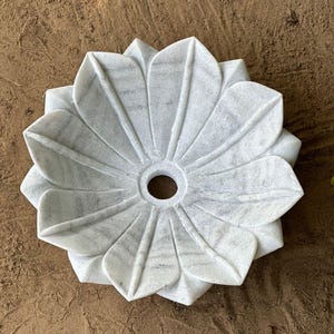 May include: A white marble decorative bowl shaped like a flower. The bowl has a central hole and is made of gray and white veined stone. The bowl is sitting on a brown surface.