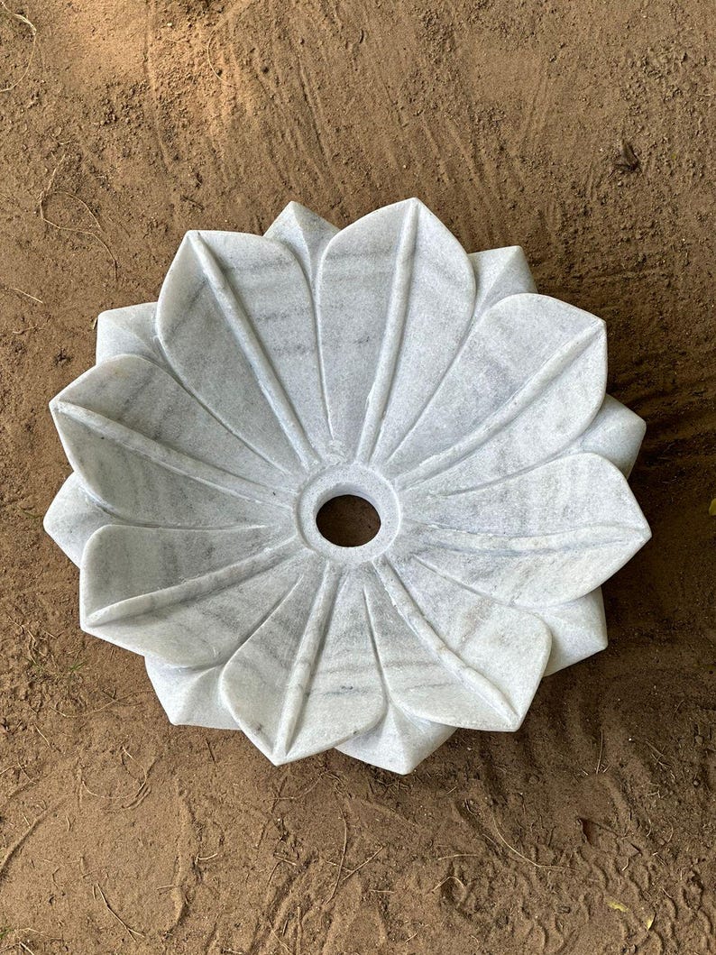 May include: A white marble decorative bowl shaped like a flower. The bowl has a central hole and is made of gray and white veined stone. The bowl is sitting on a brown surface.