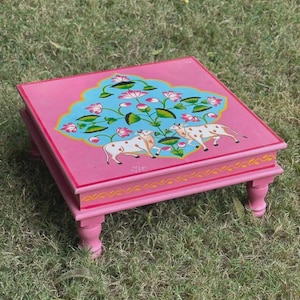 Op de afbeelding: Een roze, vierkante houten tafel met vier korte poten. De bovenkant is beschilderd met een blauw bloemmotief, twee witte koeien met bruine vlekken en roze lotusbloemen. De tafel staat op groen gras.