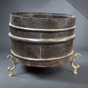 Pode incluir: Um recipiente cilíndrico de metal escuro e desgastado com duas faixas horizontais em relevo que circundam o corpo. O recipiente é suportado por três pés metálicos claros e ornamentados com um design ramificado em forma de garra.