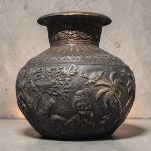 May include: A dark metal vase with intricate relief carvings. The vase features detailed scenes of trees, figures, and birds. The top and rim have a contrasting bronze color, adding to the antique aesthetic. The vase has a wide body and a flared rim.