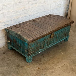 May include: A large, antique wooden trunk with a distressed teal finish and a weathered, brown metal top. The trunk has a rectangular shape with a curved lid and is raised on short legs. The metal top is secured with metal hinges and clasps.