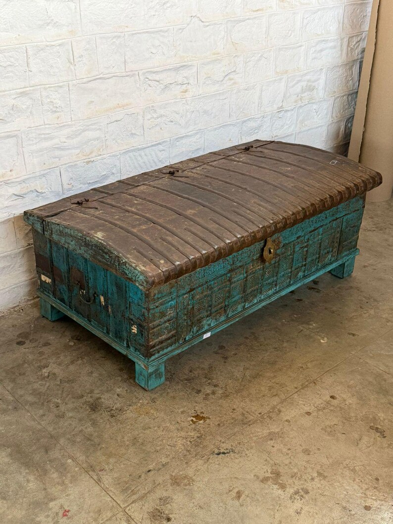 May include: A large, antique wooden trunk with a distressed teal finish and a weathered, brown metal top. The trunk has a rectangular shape with a curved lid and is raised on short legs. The metal top is secured with metal hinges and clasps.