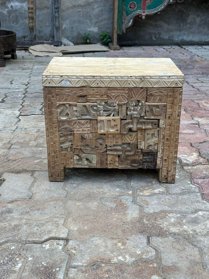 Op de afbeelding: Een handgemaakte houten bijzettafel met een lichtgekleurd blad en ingewikkeld gesneden voorpanelen. De panelen hebben een moza&iuml;ek van gesneden letters en vormen, wat een unieke, rustieke esthetiek cre&euml;ert. De tafel staat op vier korte poten.