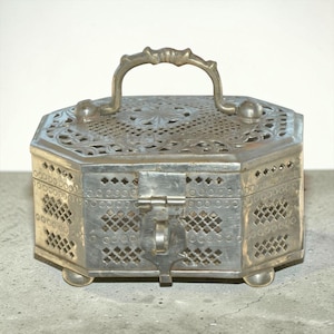May include: A silver-colored, hexagonal metal box with a decorative handle and latch. The box features intricate cut-out patterns and rests on small, rounded feet. The top has a floral design, and the handle is ornate.