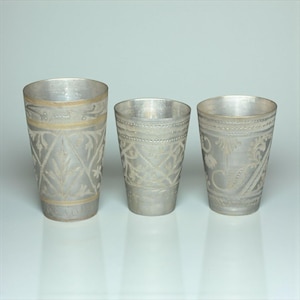 May include: Three silver-colored drinking glasses with intricate etched floral and geometric patterns. The glasses vary in size and design, with one featuring a gold-colored band near the rim. A decorative set.
