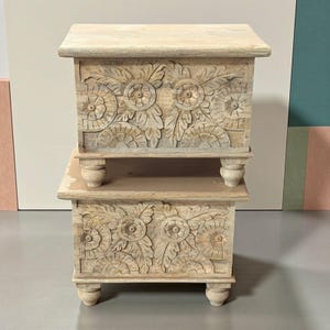 May include: Two stacked, light-colored wooden chests with intricate floral carvings. Each chest has a rectangular shape, four rounded legs, and a flat top. The carvings feature detailed flower and leaf patterns.