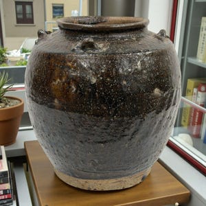 Könnte beinhalten: Ein großes, rundes Keramikgefäß mit dunkler brauner Glasur. Das Gefäß hat eine weite Öffnung, zwei kleine Griffe und eine strukturierte Oberfläche. Der untere Teil des Gefäßes ist dunkler braun. Es steht auf einer Holzoberfläche.