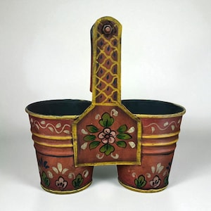 Puede incluir: Un caddy decorativo de metal pintado a mano con dos compartimentos y un asa. El caddy está pintado en tonos de rojo, amarillo y verde, con diseños florales y geométricos. El asa presenta un patrón de celosía.