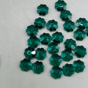May include: A collection of 20 teal green flower-shaped rhinestones. The rhinestones are scattered on a light gray surface.