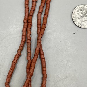 May include: Strands of reddish-brown beads, each with a cylindrical shape, are displayed against a light gray background. A U.S. quarter coin is visible, providing a size reference. The beads appear to be strung together.
