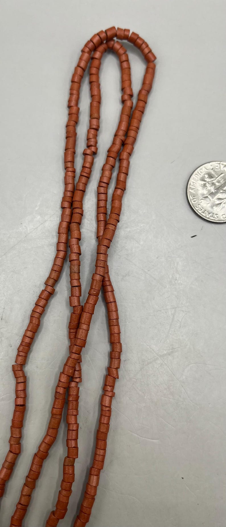 May include: Strands of reddish-brown beads, each with a cylindrical shape, are displayed against a light gray background. A U.S. quarter coin is visible, providing a size reference. The beads appear to be strung together.