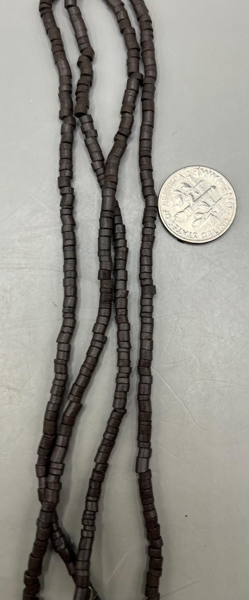 May include: Dark brown wooden bead strands with a dime for size comparison. The beads are cylindrical and strung together, creating a necklace or bracelet component. The beads are a deep, earthy tone.