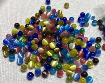 May include: A collection of colorful, round beads in various sizes and shades. The beads are a mix of colors, including blue, green, yellow, pink, and brown. Some beads have a cat's eye effect. Ideal for jewelry making and crafts.