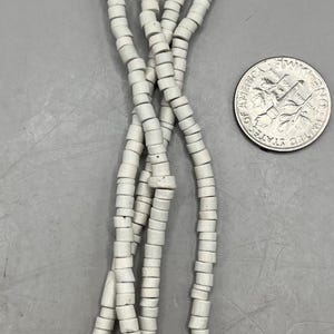 May include: Strands of off-white, cylindrical beads are displayed against a gray background, with a U.S. dime for size comparison. The beads appear to be made of a ceramic or stone material, and are strung together.