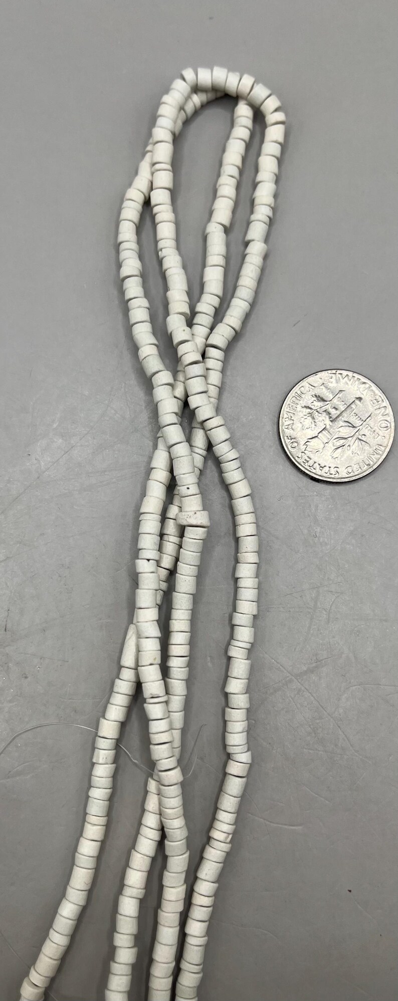 May include: Strands of off-white, cylindrical beads are displayed against a gray background, with a U.S. dime for size comparison. The beads appear to be made of a ceramic or stone material, and are strung together.