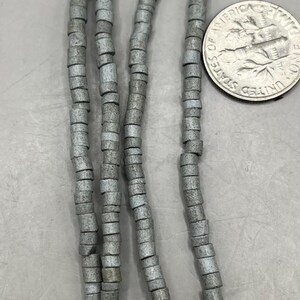 May include: Strands of gray, cylindrical beads. The beads are strung together, forming multiple strands. A US dime is next to the beads for size comparison. The beads appear to be made of stone or a similar material.
