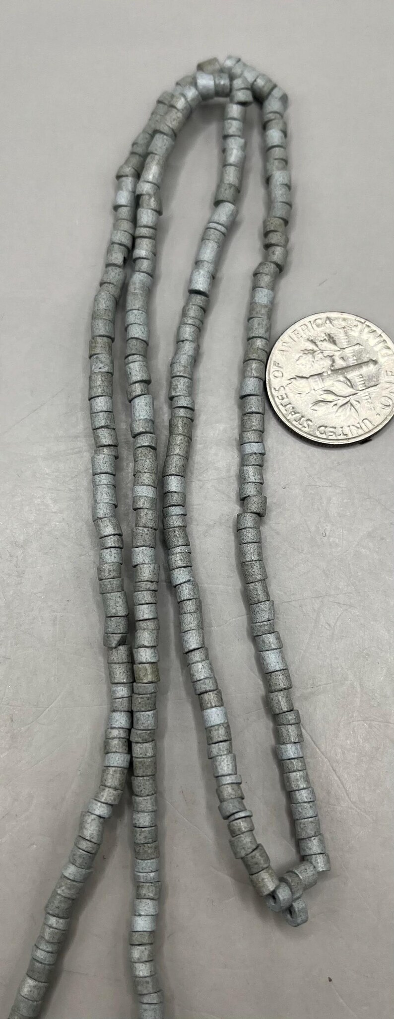 May include: Strands of gray, cylindrical beads. The beads are strung together, forming multiple strands. A US dime is next to the beads for size comparison. The beads appear to be made of stone or a similar material.