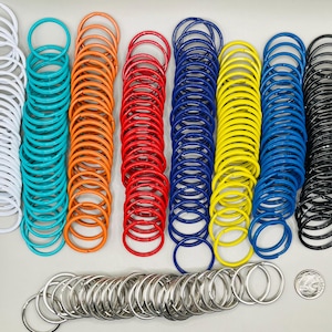 May include: Assortment of colorful metal key rings in white, turquoise, orange, red, blue, yellow, and black. A pile of silver key rings is in the foreground.