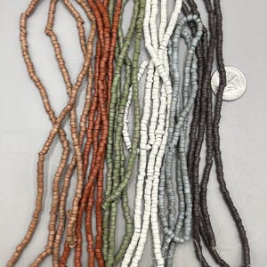 May include: A collection of beaded strands in various colors, including light brown, terracotta, olive green, white, gray, and dark brown. The beads are small, cylindrical, and strung together, creating a textured appearance. The beads are likely used for jewelry making.