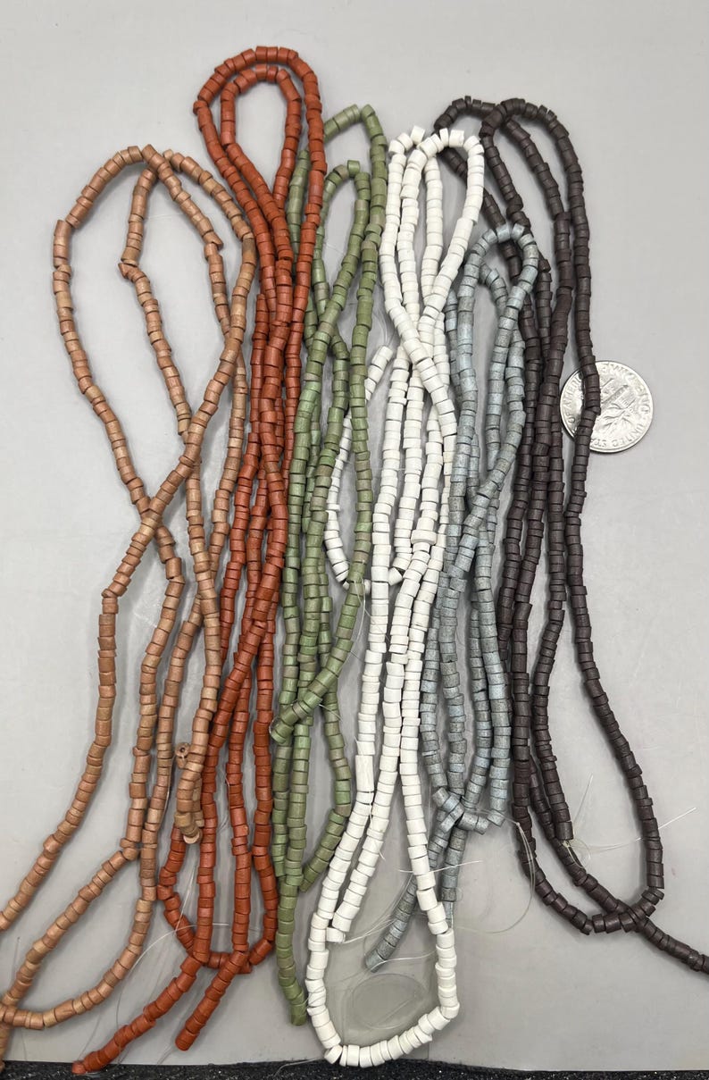 May include: A collection of beaded strands in various colors, including light brown, terracotta, olive green, white, gray, and dark brown. The beads are small, cylindrical, and strung together, creating a textured appearance. The beads are likely used for jewelry making.