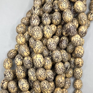 May include: Close-up of a collection of beige-colored beads with a dark brown, web-like pattern. The beads are oval-shaped and appear to be strung together. The image is well-lit, showcasing the texture and detail of the beads, which could be used for jewelry making.