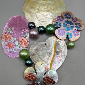 May include: A collection of iridescent shell pendants and beads. The pendants are in various shapes, including a flower, heart, and oval, with floral and geometric designs. The beads are in shades of green, black, and pink.