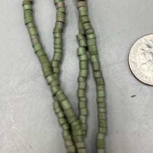 May include: Strands of olive green beads. The beads are cylindrical and strung together, creating a necklace or bracelet. The beads have a slightly weathered appearance. A US quarter is in the background.