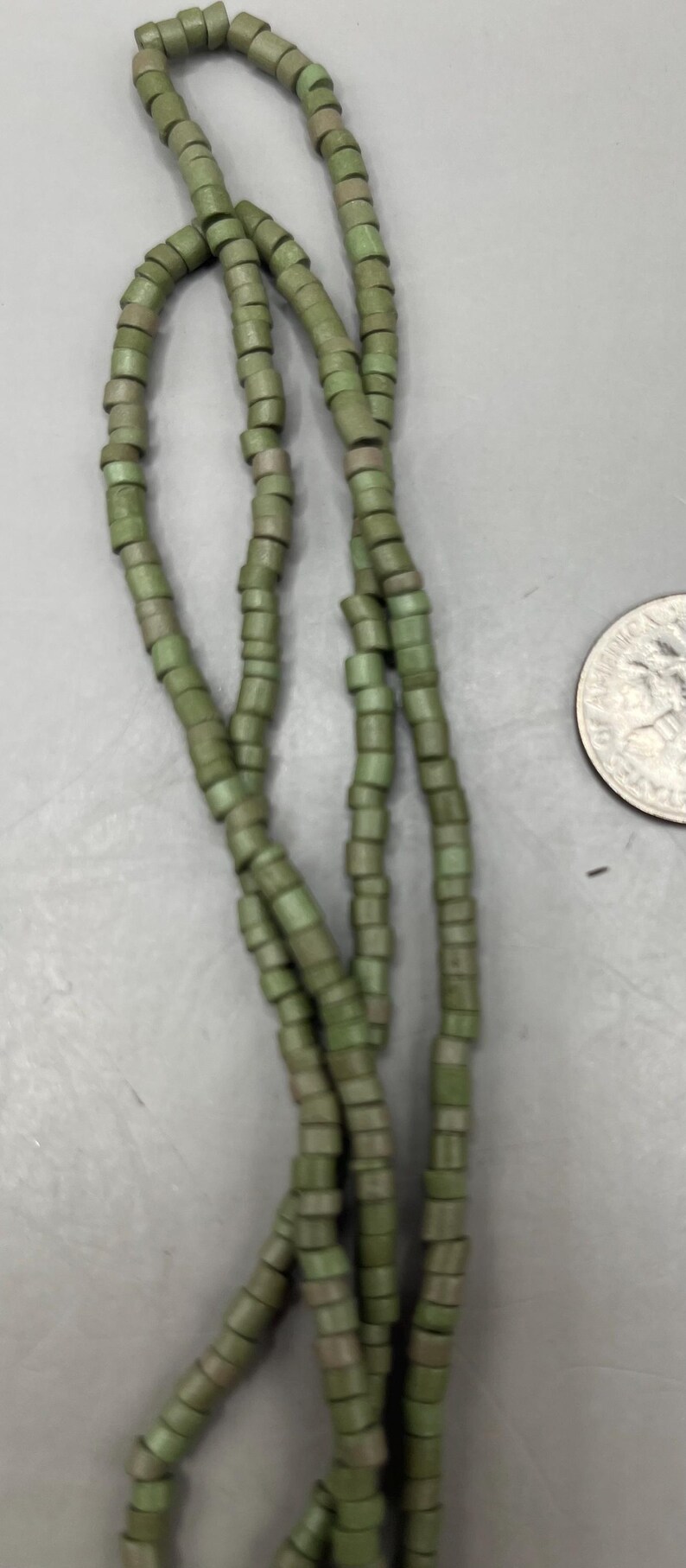 May include: Strands of olive green beads. The beads are cylindrical and strung together, creating a necklace or bracelet. The beads have a slightly weathered appearance. A US quarter is in the background.