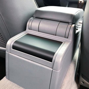 May include: Gray car center console with a black padded lid and a gray armrest. The console has a storage compartment with a sliding lid.