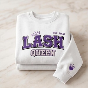 May include: White sweatshirt with the text "LASH QUEEN" in purple and white varsity-style lettering, with a crown above the text. The sleeve has a small crown and heart design. The sweatshirt is folded on a marble surface.