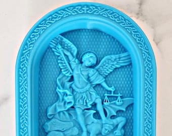 May include: A blue 3D printed figurine of an angel with wings, holding a scale and standing on a demon. The figurine is set in a blue arched frame with a decorative border.