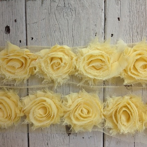 May include: A yellow fabric trim with a row of ruffled fabric flowers. The trim is on a white wooden background.