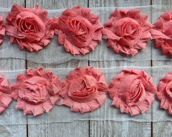 May include: A close-up of two rows of pink fabric flowers sewn onto a white mesh ribbon. The flowers are ruffled and have a delicate, feminine look.