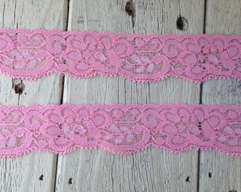 May include: Pink lace trim with a delicate floral pattern. The lace is shown on a wooden surface.
