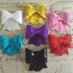 May include: Six different colored bows with sequins. The bows are in white, yellow, pink, purple, blue, and black.
