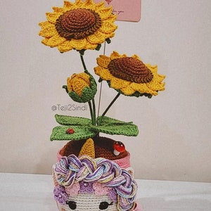 以下が含まれることがあります： ピンクのユニコーン型のプランターに、3つの黄色い花と緑の葉を持つ、かぎ針編みのひまわり植物。プランターは編まれたたてがみと可愛い顔をしています。植物の後ろにはピンクのカードに「あなたへ」の文字が書かれています。
