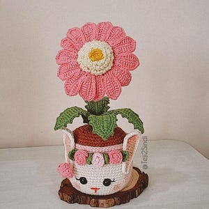 Puede incluir: Una maceta de ganchillo con una flor rosa y una cara de conejo blanco. La maceta está sentada sobre una rodaja de madera.