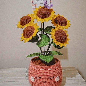 Può includere: Un bouquet di girasoli all'uncinetto in un vaso rosa con un viso sorridente. Il vaso ha una base marrone e cuciture bianche. I girasoli sono marroni e gialli con foglie verdi. Il testo sulla carta dice "Las Flores son la herramienta mas eficaz de la naturaleza para que te recuerdes que la vida es bella."