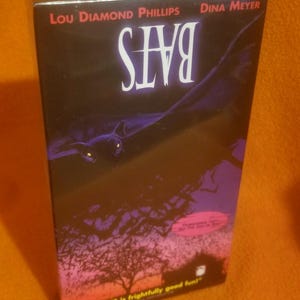 Peut inclure: Une jaquette de cassette VHS pour le film "Bats" avec Lou Diamond Phillips et Dina Meyer. La jaquette présente une chauve-souris noire volant sur un fond de ciel violet foncé avec un slogan qui dit "Bats is frightfully good fun!"