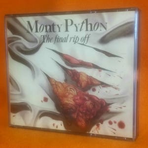 May include: A Monty Python The Final Rip Off music CD case featuring a white fabric background with three diagonal claw marks revealing red, textured, fleshy layers underneath, accompanied by scattered red blood-like droplets on the surface.