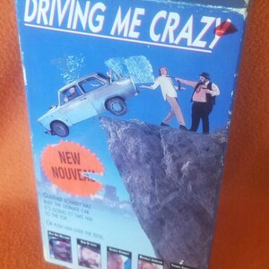 May include: A VHS video cassette cover with a blue background and the title "Driving Me Crazy" in white letters. The cover features a cartoon image of a car driving off a cliff with two men hanging on to the back. The text "New Nouveau" is in a red circle on the bottom left corner of the cover. The text "Gunther Schmidt has built the ultimate car. It's going to take him to the top. On push him over the edge." is in black text on the bottom of the cover.