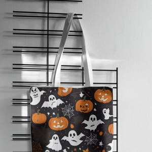 May include: A Halloween-themed tote bag with white handles. The black fabric is patterned with orange pumpkins, white ghosts, and spiderwebs. The bag is hanging on a black metal rack against a white wall.