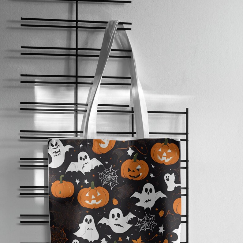 May include: A Halloween-themed tote bag with white handles. The black fabric is patterned with orange pumpkins, white ghosts, and spiderwebs. The bag is hanging on a black metal rack against a white wall.