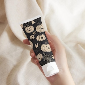 May include: A black tube with a white cap, decorated with a repeating pattern of beige jack-o'-lanterns, ghosts, and spiderwebs. The tube is held against a neutral background, suggesting a Halloween-themed product.