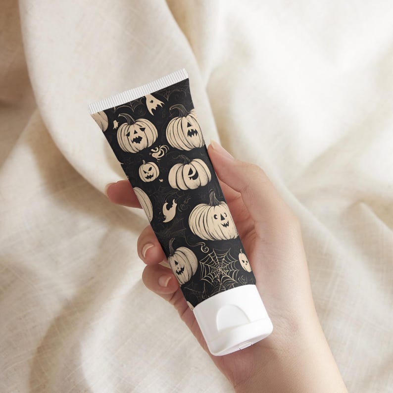 May include: A black tube with a white cap, decorated with a repeating pattern of beige jack-o'-lanterns, ghosts, and spiderwebs. The tube is held against a neutral background, suggesting a Halloween-themed product.