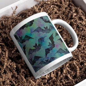 May include: White ceramic mug featuring a repeating pattern of black bats against a teal and purple watercolor background. The mug has a curved handle and is presented in a box with brown paper filler.