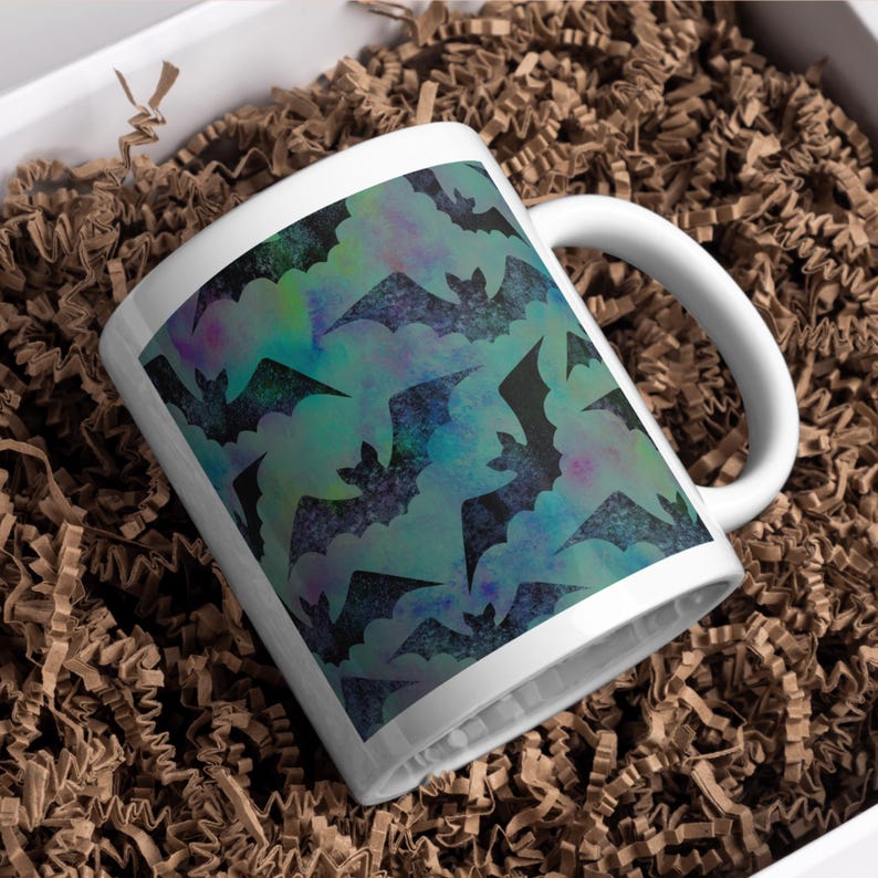 May include: White ceramic mug featuring a repeating pattern of black bats against a teal and purple watercolor background. The mug has a curved handle and is presented in a box with brown paper filler.